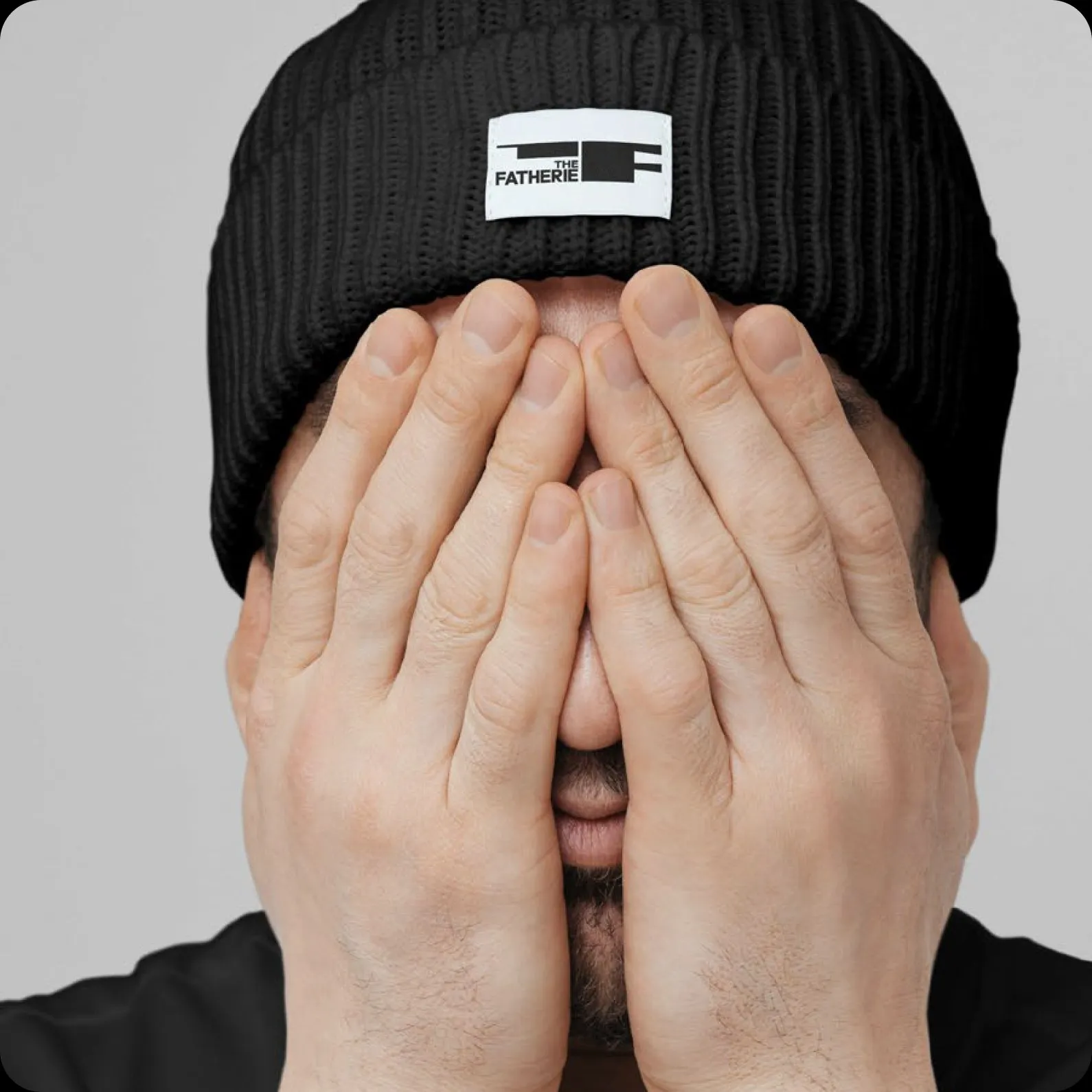 A man in a black Fatherie beanie covering his face with both hands