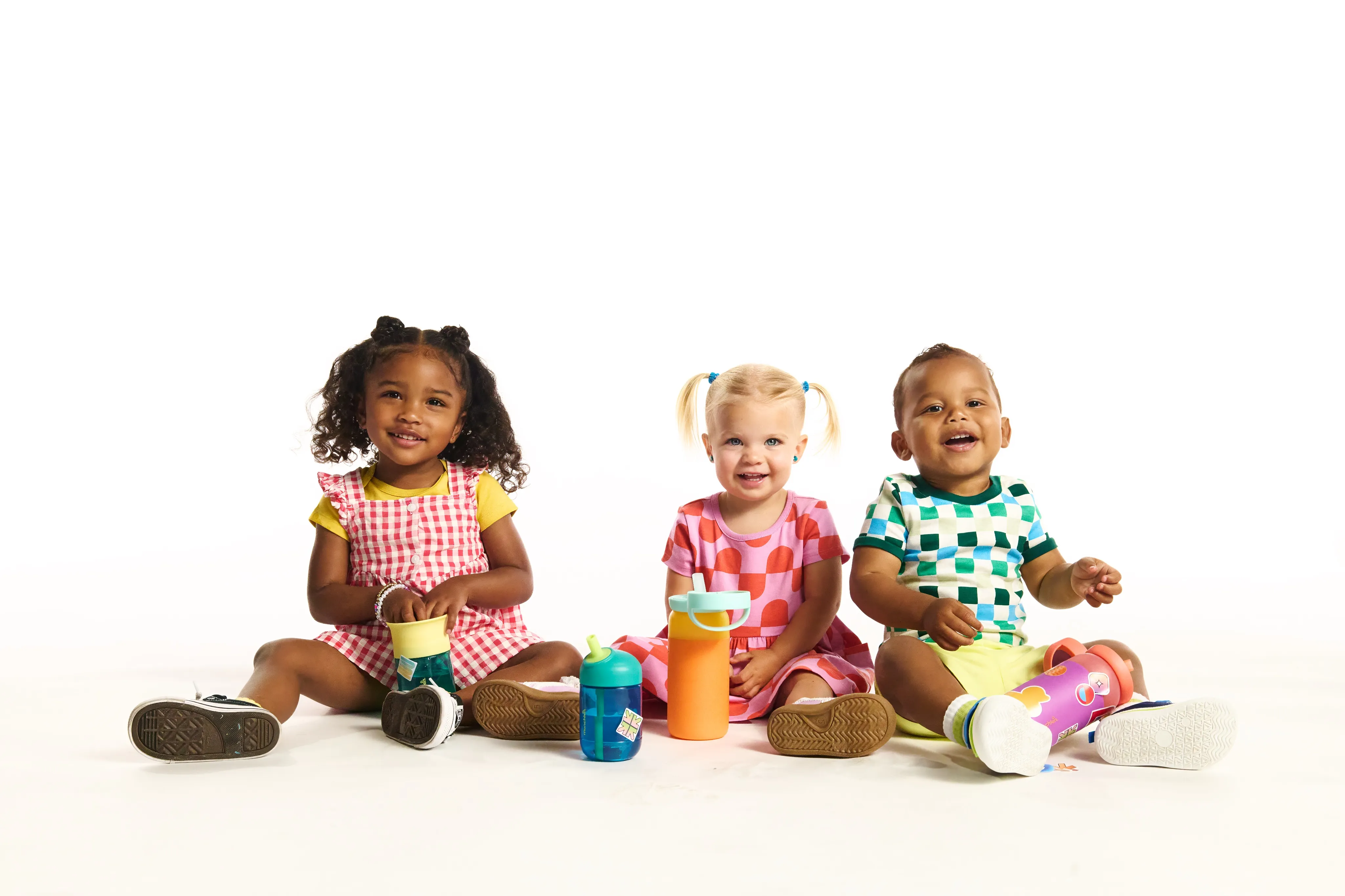 Three toddlers sitting together with Munchkin cups and bottles